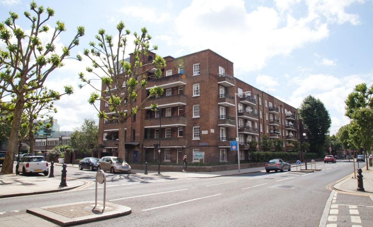 A street view of a brick-built apartment block with multiple balconies, surrounded by a well-maintained urban environment. The road in front has cars parked along the side, a pedestrian crossing, and trees with trimmed branches lining the sidewalk.