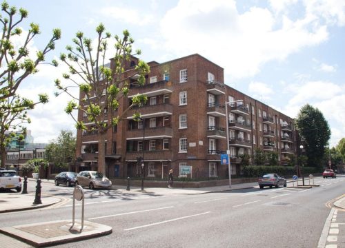 A street view of a brick-built apartment block with multiple balconies, surrounded by a well-maintained urban environment. The road in front has cars parked along the side, a pedestrian crossing, and trees with trimmed branches lining the sidewalk.