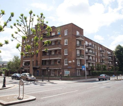 A street view of a brick-built apartment block with multiple balconies, surrounded by a well-maintained urban environment. The road in front has cars parked along the side, a pedestrian crossing, and trees with trimmed branches lining the sidewalk.