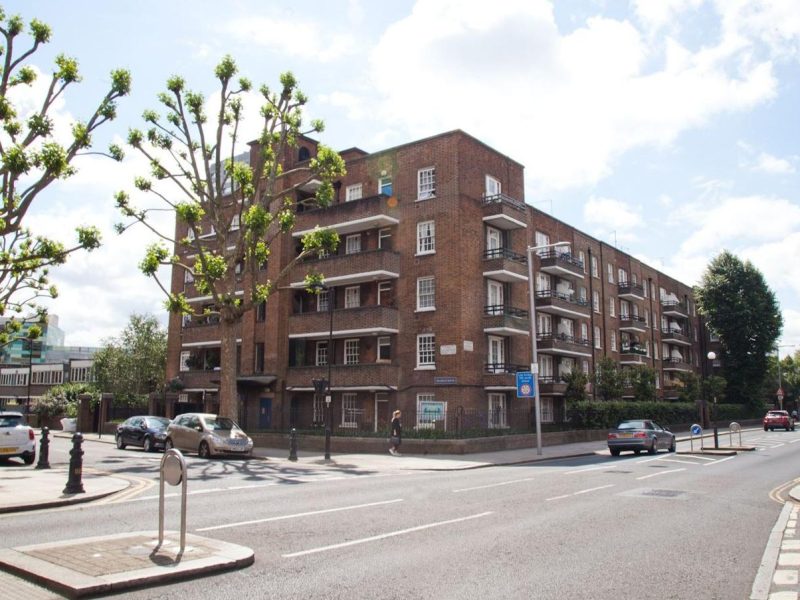 A street view of a brick-built apartment block with multiple balconies, surrounded by a well-maintained urban environment. The road in front has cars parked along the side, a pedestrian crossing, and trees with trimmed branches lining the sidewalk.