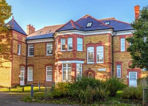 Wide-angle view of a grand Victorian brick building with red accents, multiple large windows, blue roof tiles, and lush landscaping in the foreground, taken on a sunny day.