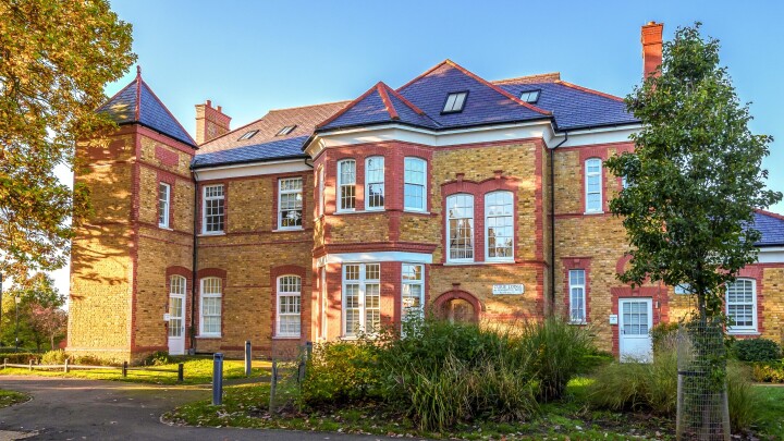 Wide-angle view of a grand Victorian brick building with red accents, multiple large windows, blue roof tiles, and lush landscaping in the foreground, taken on a sunny day.