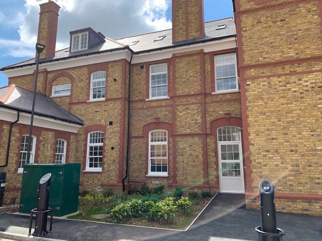 Exterior view of a renovated Victorian-style brick building with large white-framed sash windows, landscaped greenery, electric vehicle charging points, and a clear sky above.