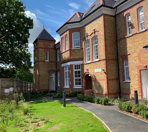 View of a Victorian brick building with a turret, pathway, lawns, and a partly cloudy sky, with well-maintained greenery surrounding the entrance.