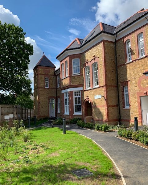 View of a Victorian brick building with a turret, pathway, lawns, and a partly cloudy sky, with well-maintained greenery surrounding the entrance.