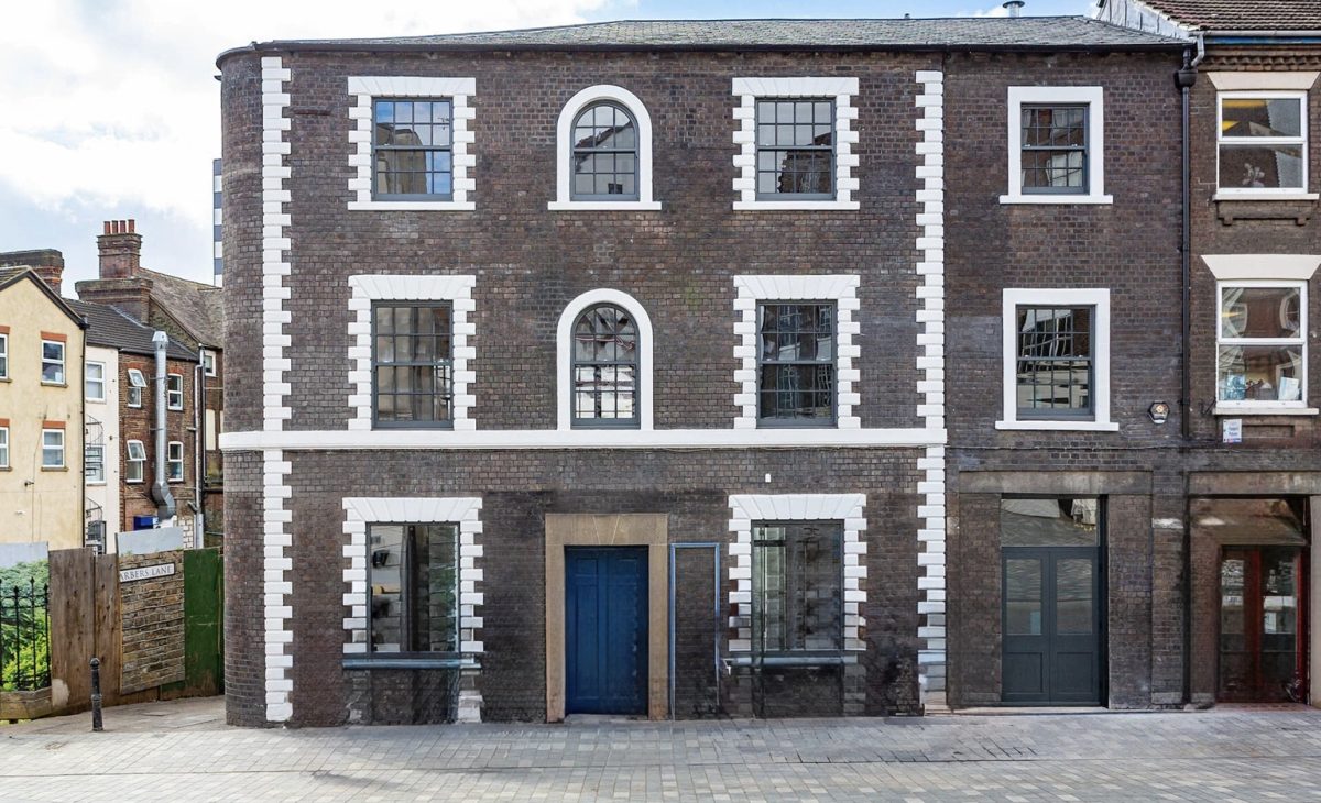 The front exterior of a three-story brick building with white-framed windows and a blue door, situated on a cobbled street with other buildings visible to the left and right.