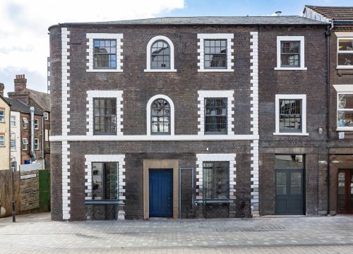 The front exterior of a three-story brick building with white-framed windows and a blue door, situated on a cobbled street with other buildings visible to the left and right.