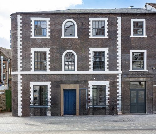 The front exterior of a three-story brick building with white-framed windows and a blue door, situated on a cobbled street with other buildings visible to the left and right.