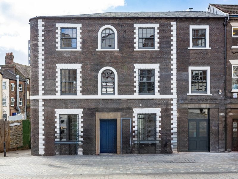 The front exterior of a three-story brick building with white-framed windows and a blue door, situated on a cobbled street with other buildings visible to the left and right.
