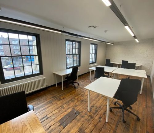 An interior view of a room with wooden floors, white walls, and large windows, furnished with several white desks and black office chairs.