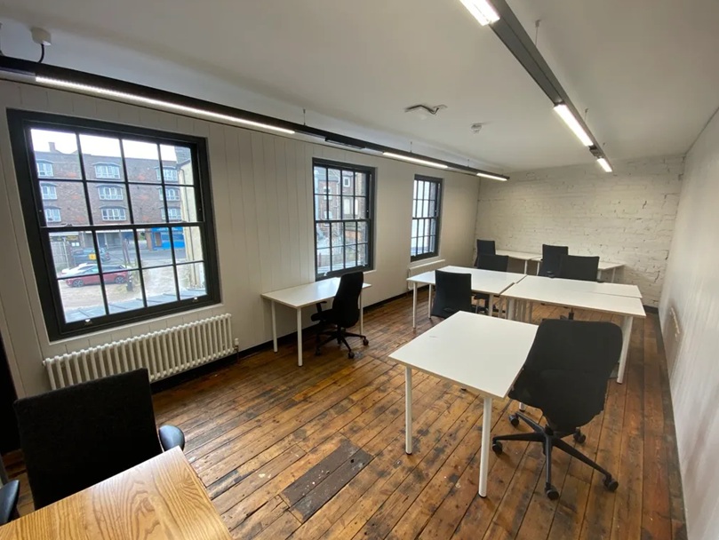 An interior view of a room with wooden floors, white walls, and large windows, furnished with several white desks and black office chairs.