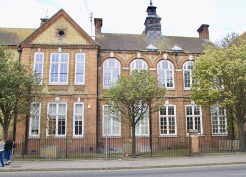 A red and yellow brick building with many large white-framed windows and a black fence in front. Two people walk on the pavement to the left, and a tree stands in the middle foreground.