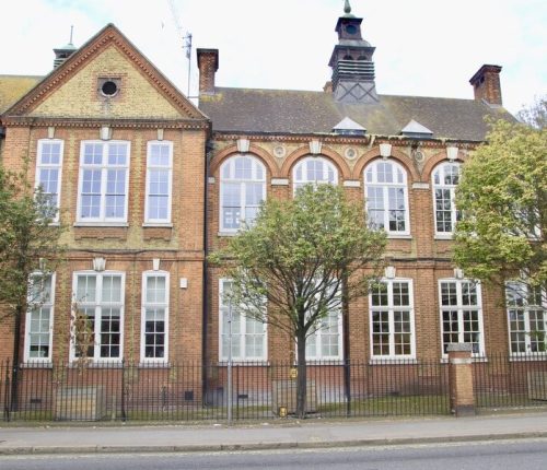 A red and yellow brick building with many large white-framed windows and a black fence in front. Two people walk on the pavement to the left, and a tree stands in the middle foreground.