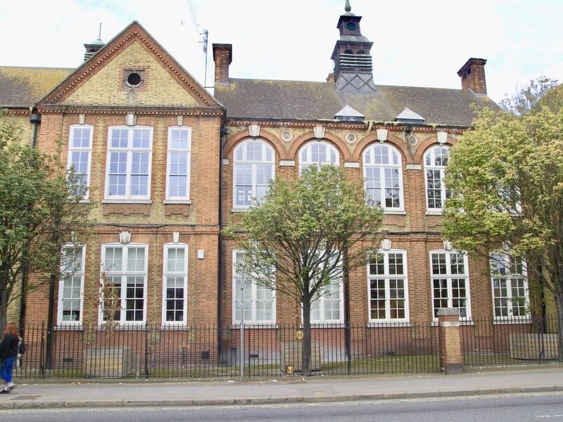 A red and yellow brick building with many large white-framed windows and a black fence in front. Two people walk on the pavement to the left, and a tree stands in the middle foreground.