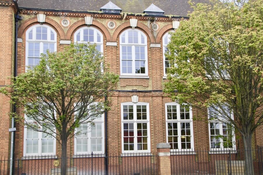 A section of a brick building featuring large arched windows on the upper floor and rectangular windows below, with two trees in front and a black fence.