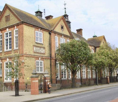 A long view of a yellow and red brick building with many large windows, a fenced front, and trees. A small archway and gate are visible on the left, and two people stand near the gate.