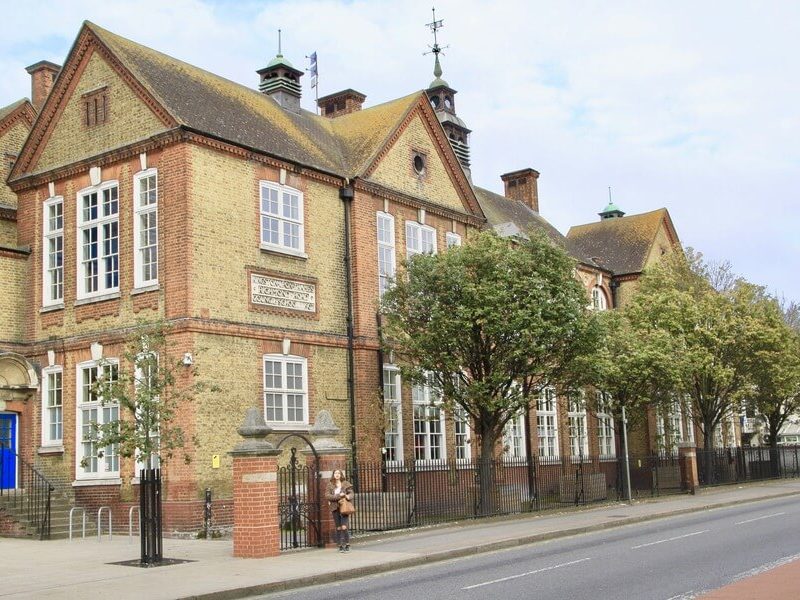 A long view of a yellow and red brick building with many large windows, a fenced front, and trees. A small archway and gate are visible on the left, and two people stand near the gate.