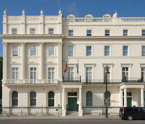 Close-up of the central part of the German Embassy in London, highlighting the main entrance, two flags, and the classical architectural details of the white facade.