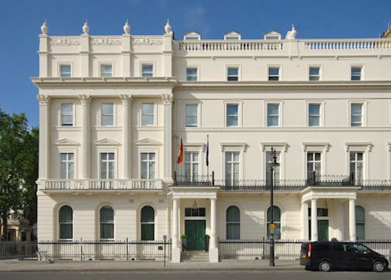 Close-up of the central part of the German Embassy in London, highlighting the main entrance, two flags, and the classical architectural details of the white facade.