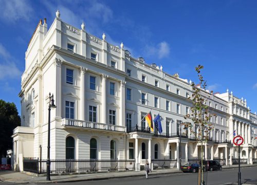 Exterior of the German Embassy in London on a sunny day, showing its white neoclassical facade and the German and EU flags flying above the entrance balcony.