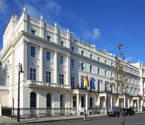 Exterior of the German Embassy in London on a sunny day, showing its white neoclassical facade and the German and EU flags flying above the entrance balcony.
