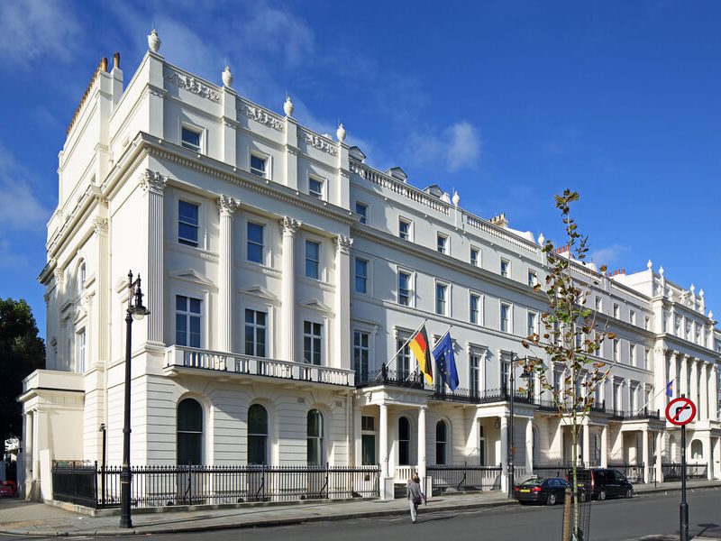 Exterior of the German Embassy in London on a sunny day, showing its white neoclassical facade and the German and EU flags flying above the entrance balcony.