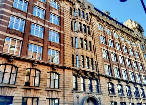 A historic building with a rich facade of alternating brown and cream bricks features arched windows and decorative elements. It stands against a clear blue sky beside a streetlight.