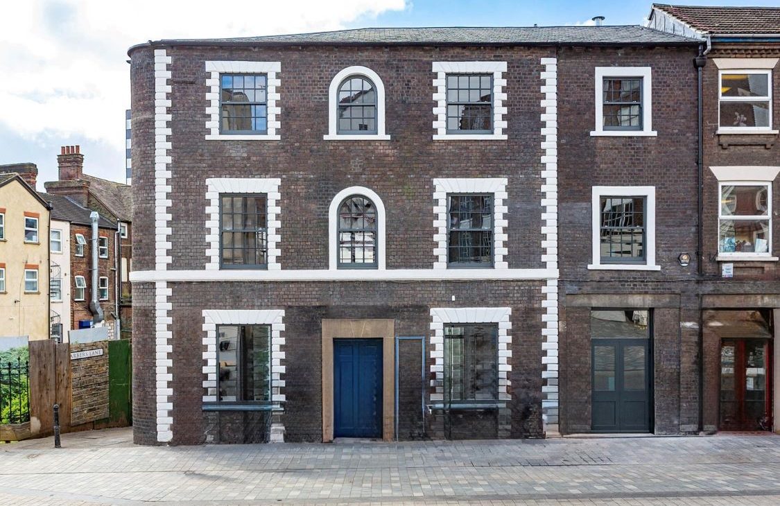 The front exterior of the three-story Luton hat factory - a brick building with white-framed windows and a blue door, situated on a cobbled street with other buildings visible to the left and right.
