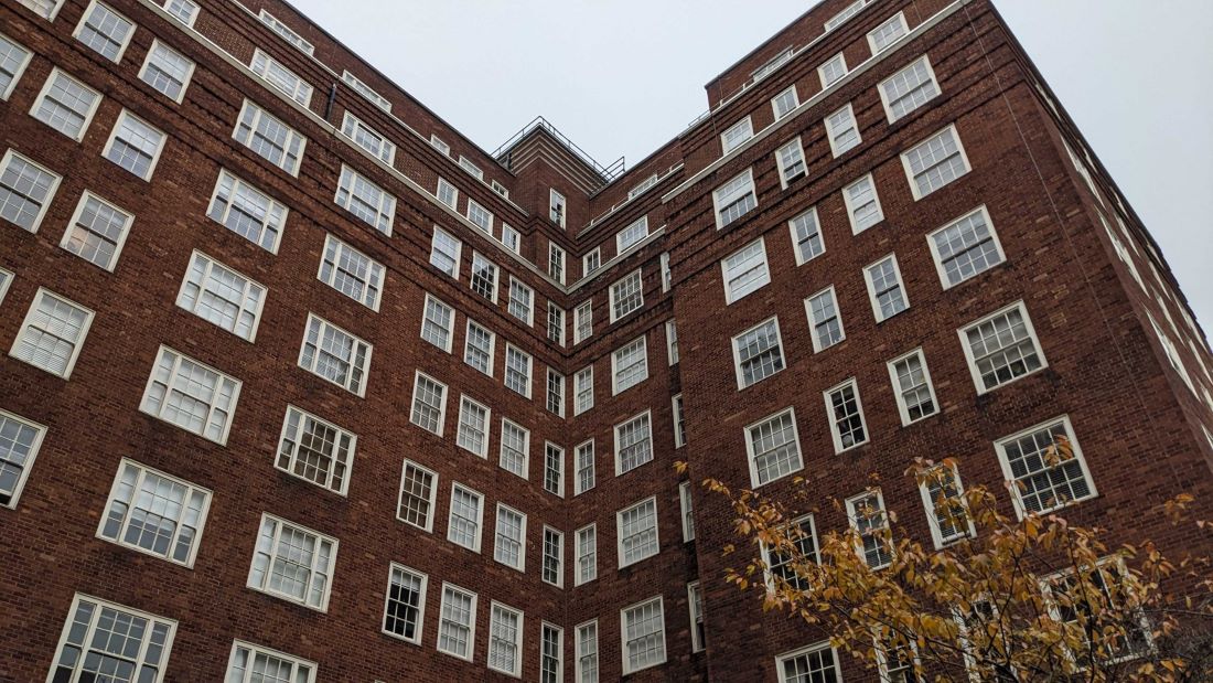 A large brick building with numerous symmetrical windows against a cloudy sky. A hint of autumn foliage is visible at the bottom.