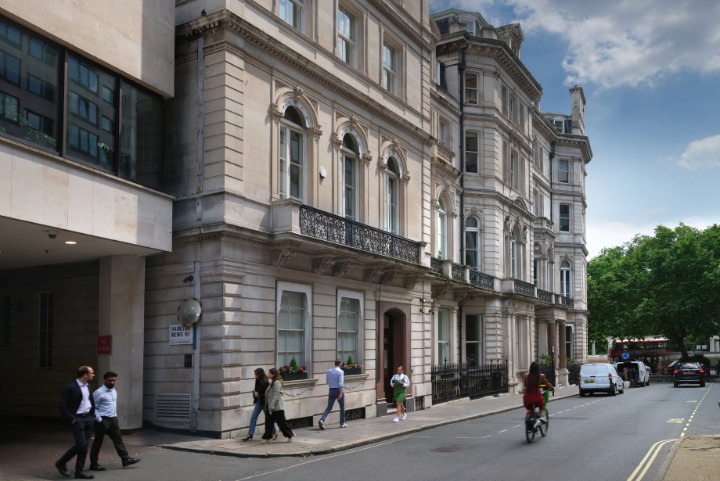 A street scene in London featuring a row of grand, multi-story neoclassical buildings with ornate stonework and wrought-iron balconies. Pedestrians walk along the sidewalk, a cyclist is in the road, and a white van and red double-decker bus are visible in the background under a partly cloudy sky.
