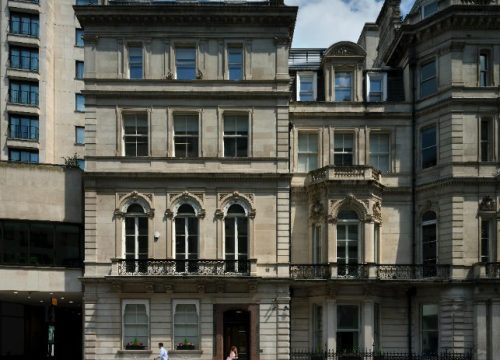 A street view of a multi-story, classical stone building in London featuring ornate architectural details, arched windows with black wrought-iron balconies, and a dark front door. Two pedestrians walk on the sidewalk in front, and a black car is parked to the right on a paved road marked with 