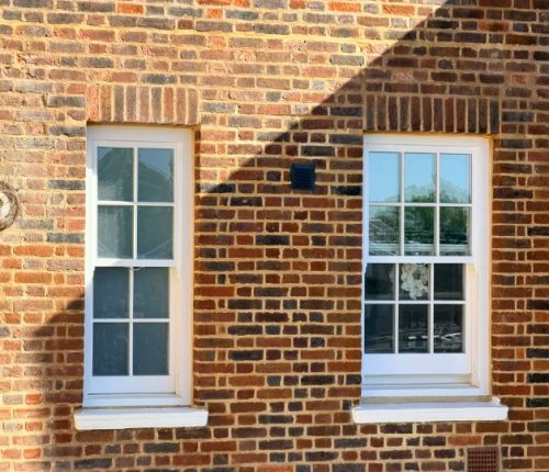 A straight-on view of two white, multi-paned wooden sash windows set into a textured brick wall. The bricks feature a mix of red, orange, and dark brown tones, laid with thick mortar. A sharp shadow from an overhanging roof cuts diagonally across the upper portion of the wall.