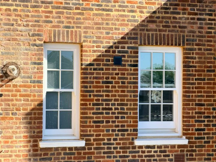 A straight-on view of two white, multi-paned wooden sash windows set into a textured brick wall. The bricks feature a mix of red, orange, and dark brown tones, laid with thick mortar. A sharp shadow from an overhanging roof cuts diagonally across the upper portion of the wall.