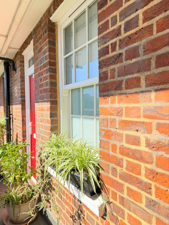 A close-up, angled view of a white multi-paned sash window on a brick building's exterior balcony. A black window box filled with vibrant green and white spider plants sits on the white sill. The brickwork shows a distinct horizontal shadow line, with darker bricks above and brighter orange-red bricks below. A red door is visible further down the walkway.
