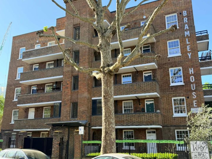 A wide exterior view of the red brick apartment complex, highlighting the balcony and window layout alongside a large mature tree in the front courtyard.
