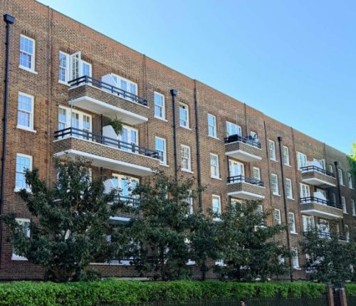 Side view of a multi-story, red brick apartment building featuring several rows of balconies with black railings and white window frames.