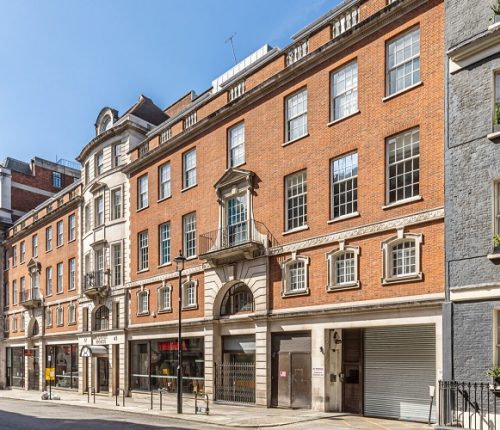 An angled, low-angle view showing a row of diverse building facades, including stone structures with classical columns, a central red brick building, and a light stone building with a small balcony.