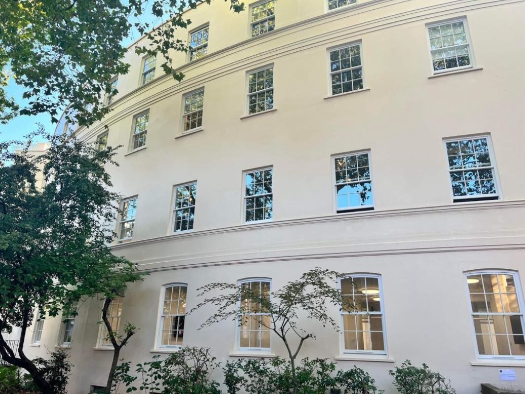 A cream-coloured building at London Business School with multiple windows reflecting trees, flanked by green foliage in daylight.