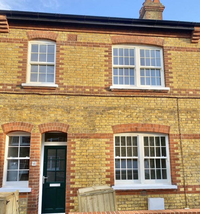 A brick wall features two arched timber windows and a central doorway. Sunlight highlights the textured yellow bricks and red brick details, enhancing the architectural character of the building.