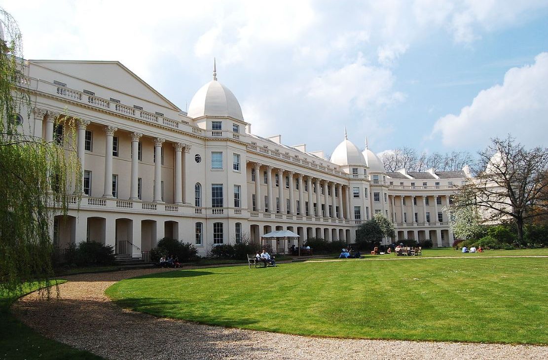 London Business School campus with white domes, surrounded by green lawns and trees, under a partly cloudy sky. People are relaxing and socialising in the grassy area.