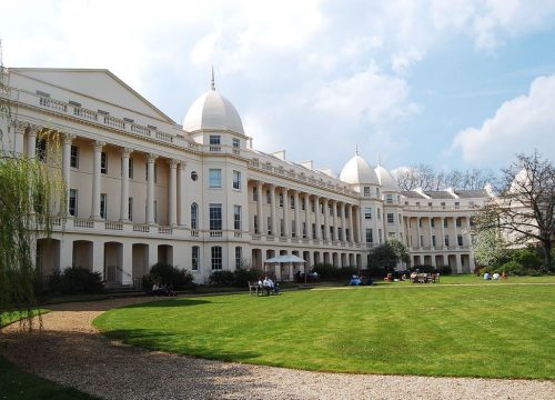 London Business School campus with white domes, surrounded by green lawns and trees, under a partly cloudy sky. People are relaxing and socialising in the grassy area.