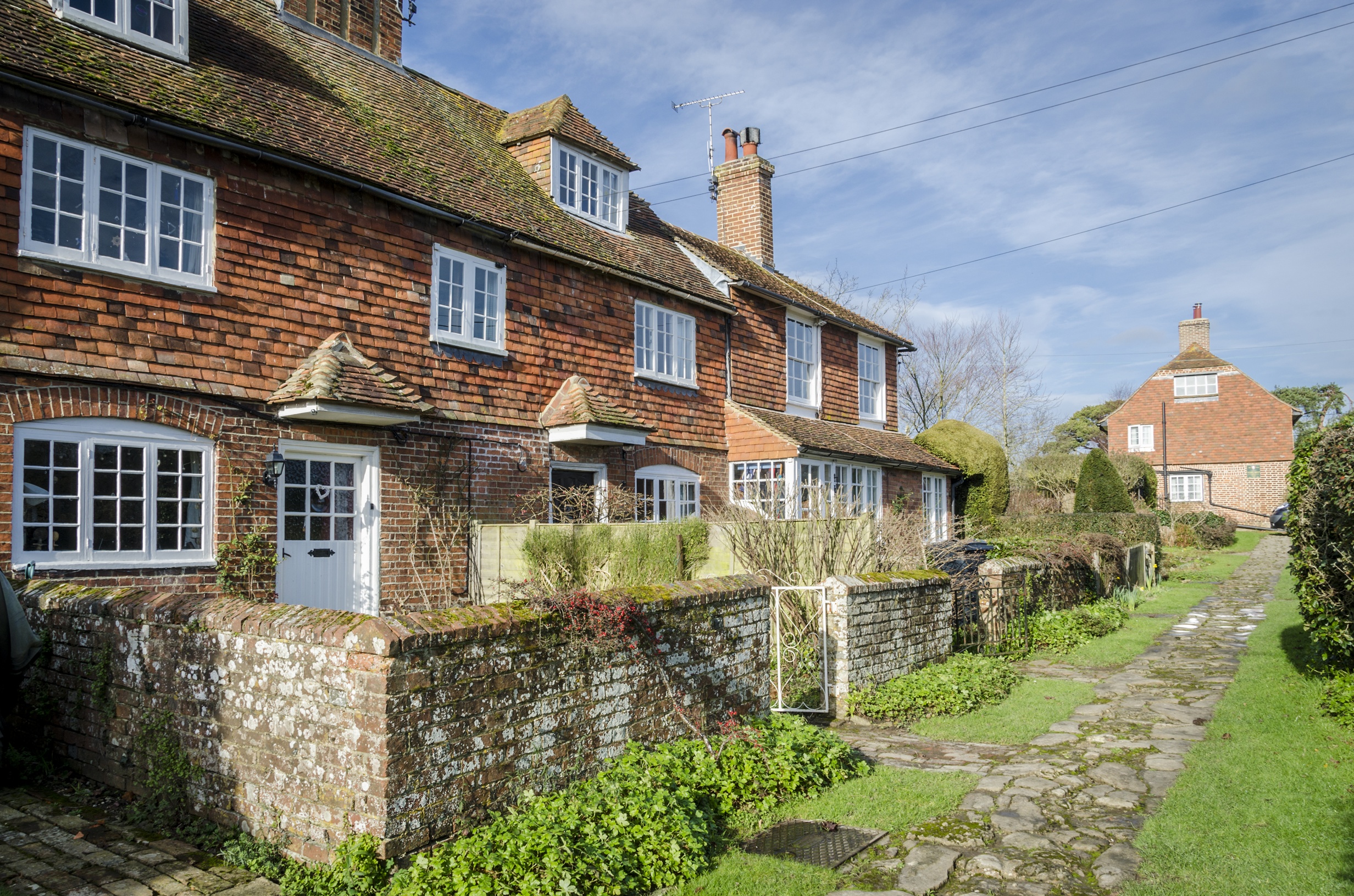 Row of traditional brick cottages with tiled roofs, low stone garden walls, and a cobblestone pathway running along a grassy area.