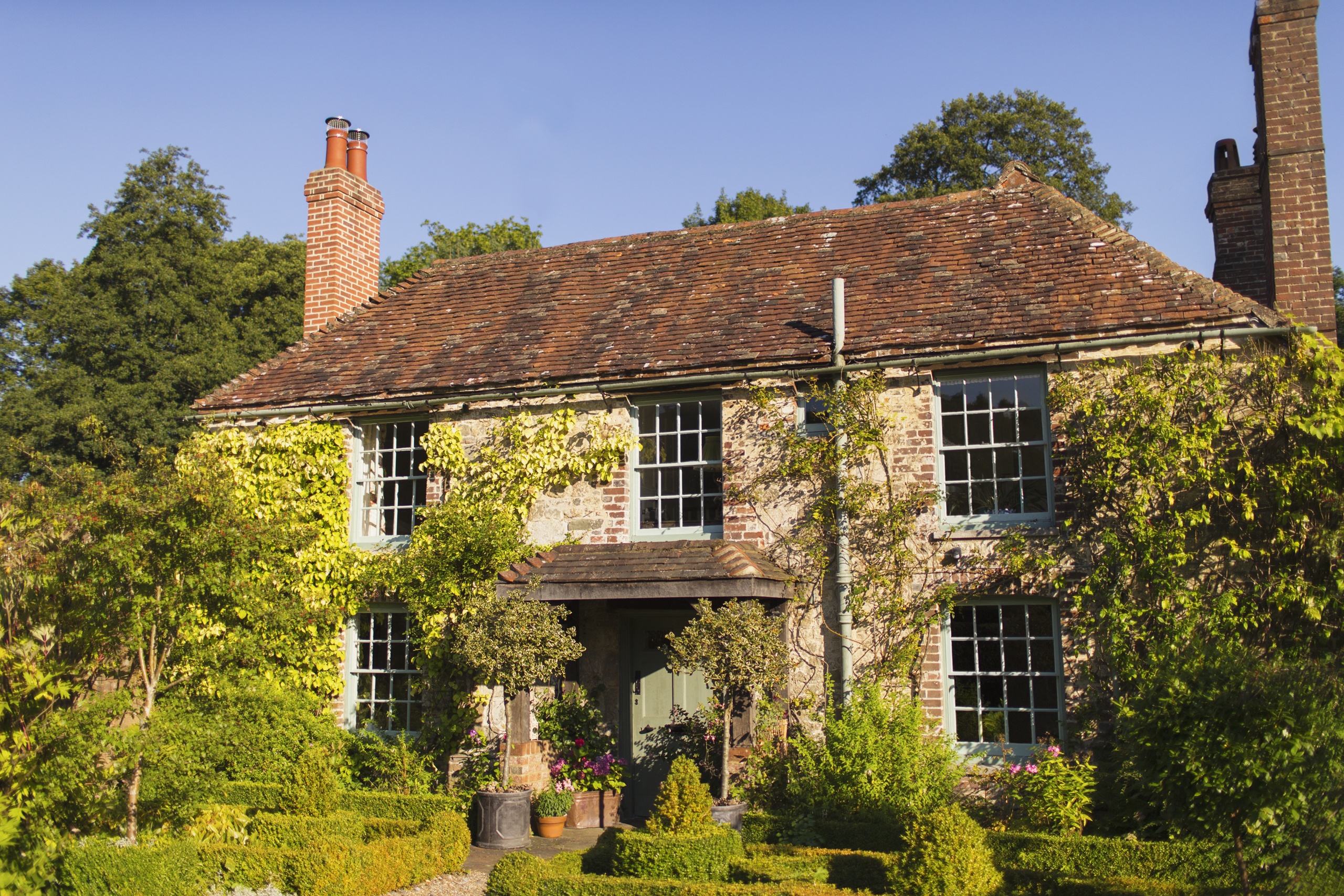 Two-story historic stone cottage with a tiled roof, tall chimneys, climbing greenery, and a manicured garden in the foreground.