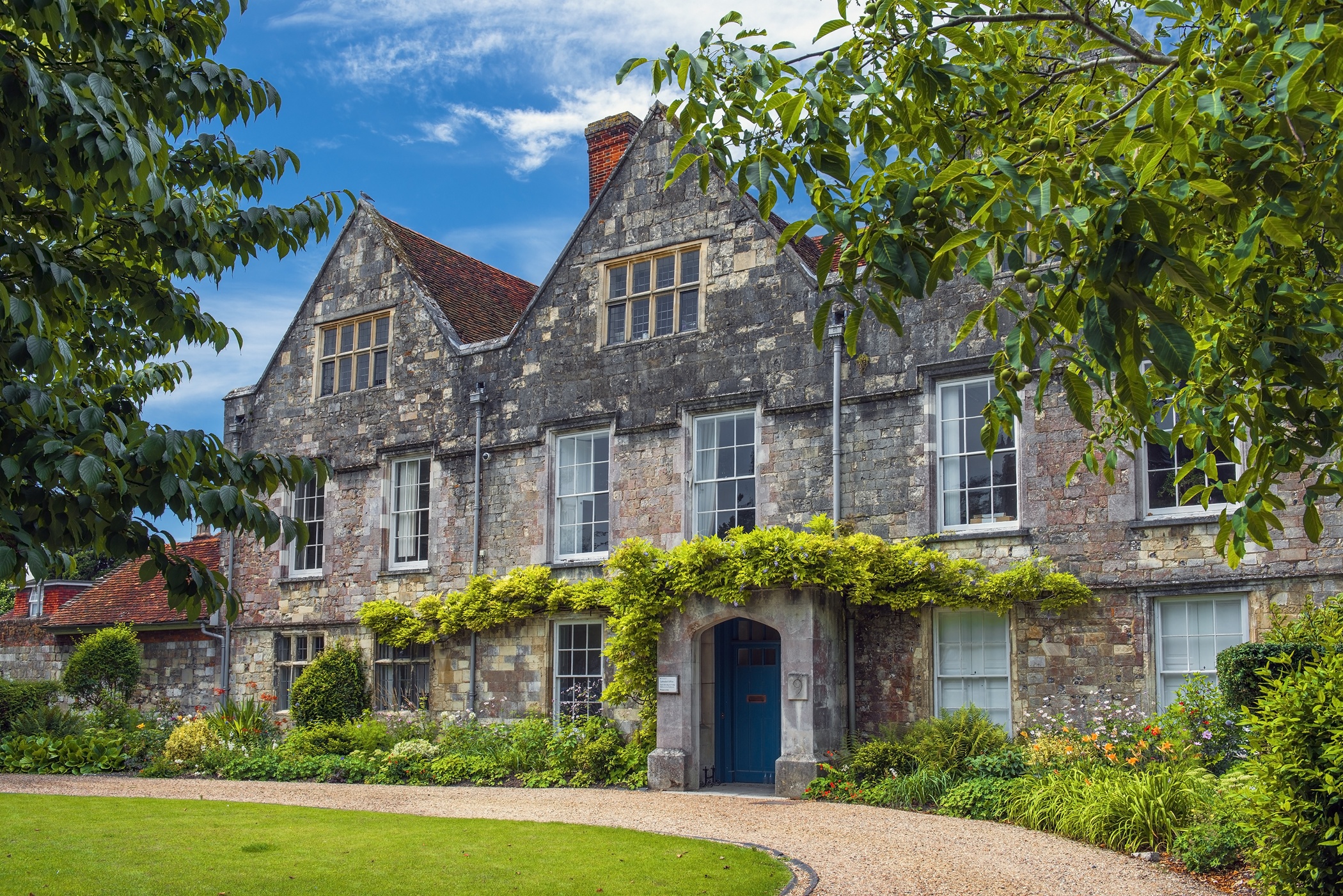 Historic historic stone manor house with gables, sash windows, and green vines framed by lush foliage and a gravel driveway.