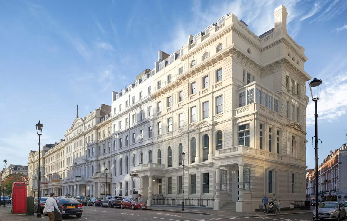 Wide street-level view of an elegant, multi-storey cream white stucco Victorian terrace building on a London street, with parked cars, a red telephone booth, and street lamps under a bright blue sky.