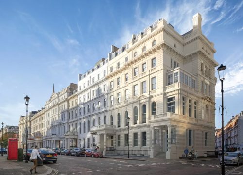 Wide street-level view of an elegant, multi-storey cream white stucco Victorian terrace building on a London street, with parked cars, a red telephone booth, and street lamps under a bright blue sky.