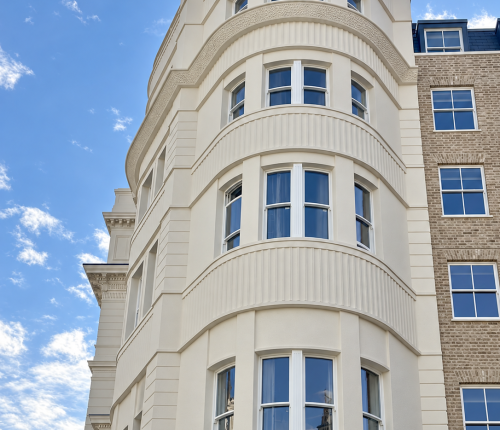 Low-angle exterior shot looking up at a curved bay window section of a newly restored off-white building facade, featuring multi-pane sash windows and classic architectural moulding against a blue sky.