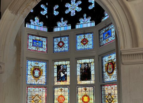 A close-up of a rectangular stained glass window pane featuring an intricate central red and gold medallion with a commemorative inscription surrounded by ornate scrollwork and a decorative border.