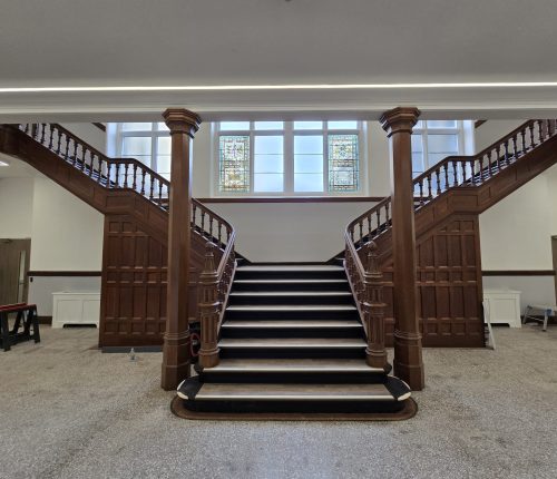 Front view of a grand wooden grand staircase splitting into two flights in a renovated atrium, with stained glass windows in the center background and polished stone flooring.