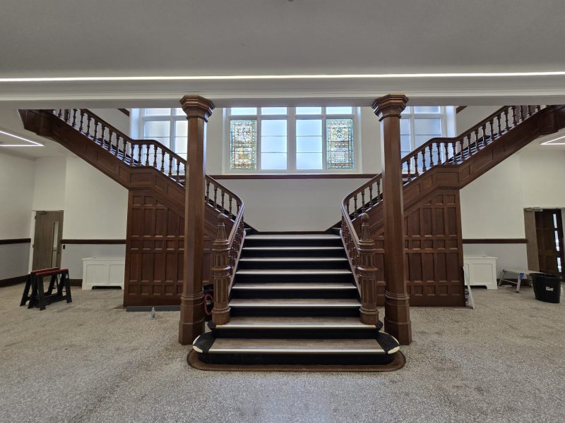 Front view of a grand wooden grand staircase splitting into two flights in a renovated atrium, with stained glass windows in the center background and polished stone flooring.